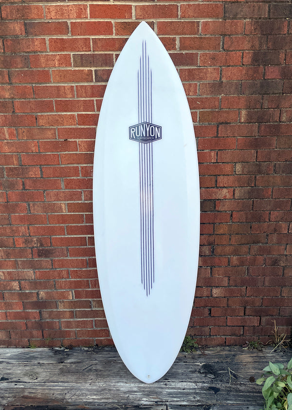 White Surfboard leaning against brick wall. Runyon Logo, 6 Stripes run from the nose, to 3 quarters of the way down the board towards the tail.