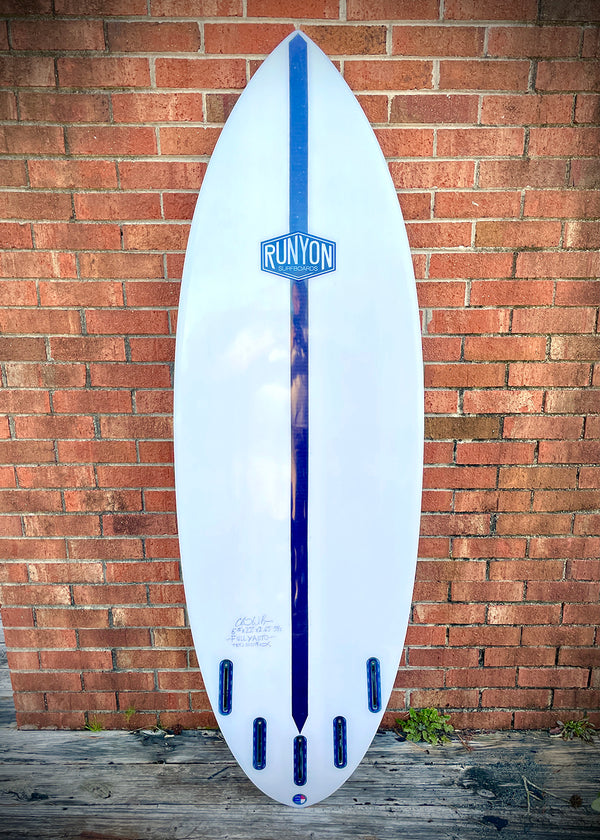 Runyon Logo, in blue. On a white surfboard, leaning against a brick wall. 4, Plus 1 fin setup, Future Fin Boxes. Wide Shortboard.