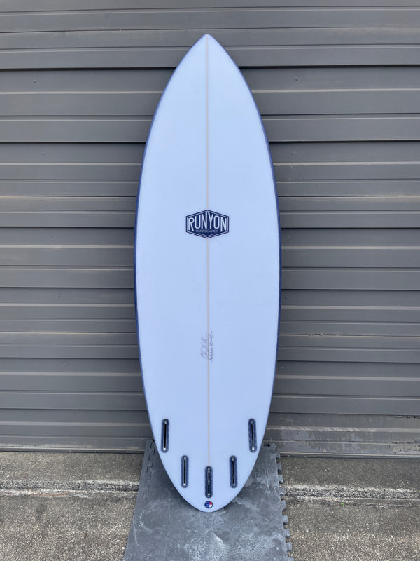 White surfboard leaning against sheet metal wall. Runyon Logo in the center. 4 +1 Future Fin Setup. Wider shortboard.