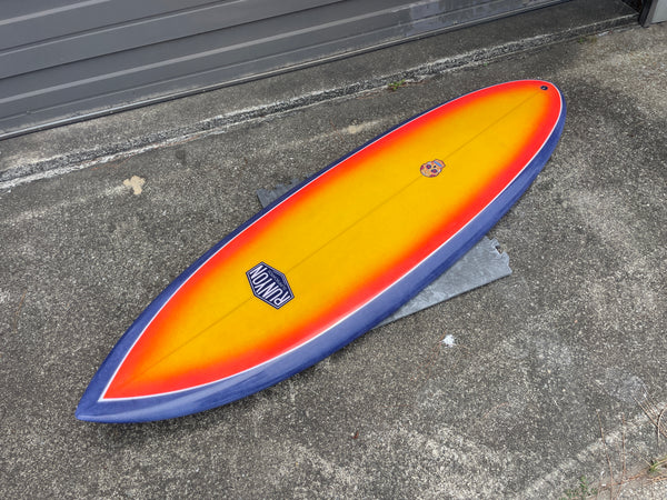 Surboard laying on cement. Blue edges, Sunset red to yellow color in the center. Runyon Logo, with skull towards the tail