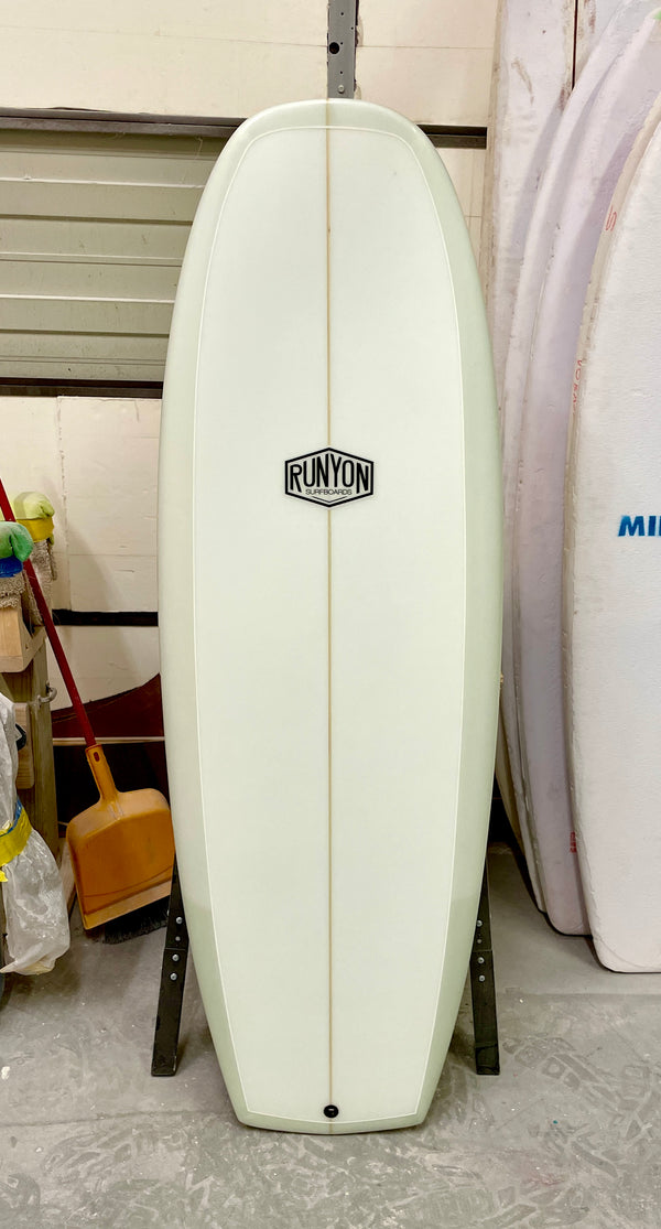 A classic-white Mini Hammer Runyon Surfboard, standing vertically in a workshop with surfboard blanks in the background.
