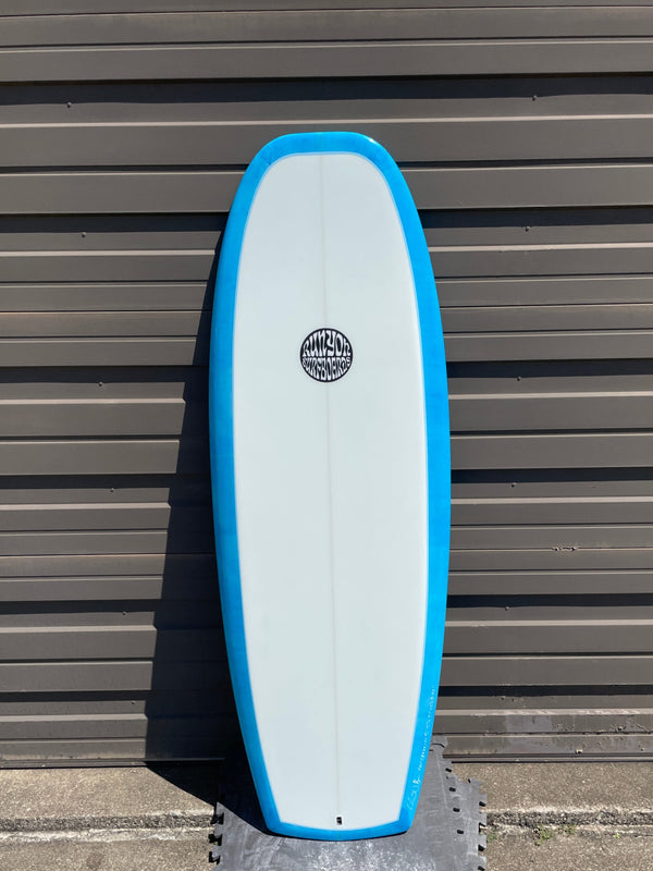 White Mini Hammer surfboard with light blue edges leaning against a sheet metal wall. Runyon Logo in the center.