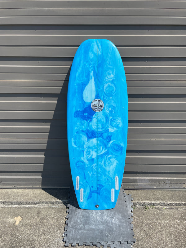 A light blue swirled pattern Mini hammer surfboard leaning against a sheet metal wall. Quad Fin Setup with White Future Fin Boxes.