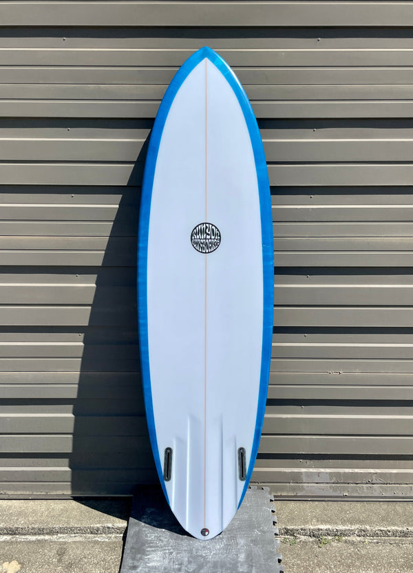 Blue Runyon "Dang Dang" Midlength Surfboard. Blue Marbling on Surfboard. Board is outside, leaning against a metal fabricate wall