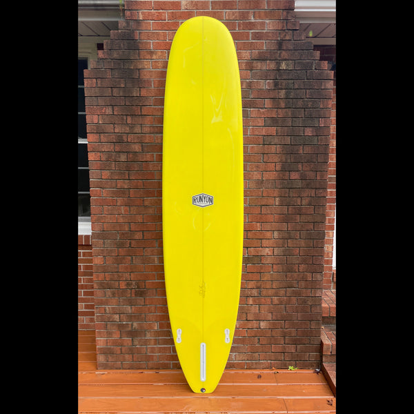 A Yellow Runyon Longboard leaning against a fireplace, outside. 3 fin setup.