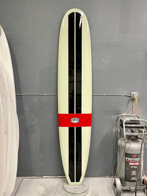 A long surfboard up against a wall, 2 vertical black stripes, 1 horizontal red band "Runyon" logo in the center. 