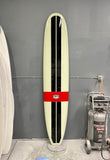 A long surfboard up against a wall, 2 vertical black stripes, 1 horizontal red band "Runyon" logo in the center. 
