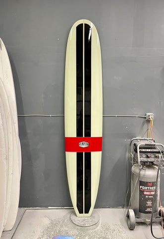A long surfboard up against a wall, 2 vertical black stripes, 1 horizontal red band "Runyon" logo in the center. 