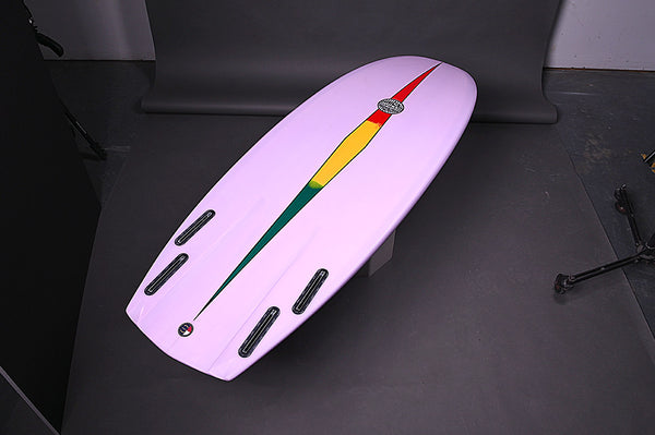 Light Purple Mini Hammer Surfboard with elongated greed, yellow, and red diamond stratching from tail to nose. Runyon logo in the center. Quad Fin Setup. Laying on its deck in a photography studio. 