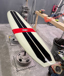 A white and black Runyon surfboard with a red stripe, sitting among various tools and equipment in what appears to be a garage or workshop.