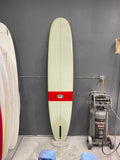 A white surfboard with a black center stripe and red tail block featuring the "RUNYON" logo, leaning against a gray tiled wall.