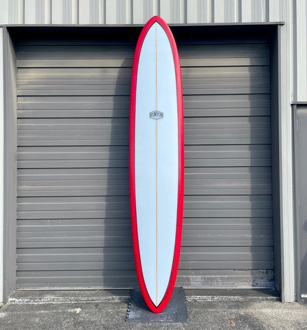 A red framed, white center, glider surfboard leaning against a metal garage door. Runyo logo in the center. 