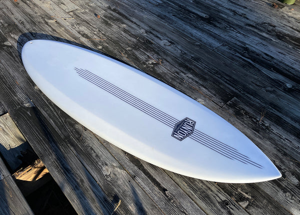 White Fully Auto surfboard, laying on wooden deck. Runyon logo towards nose, 6 stripes running vertically down the board, 