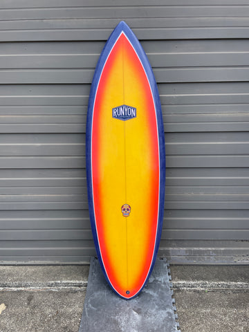 Surfboard Standing against sheet metal wall, blue edges, sunset yellow to orange gradient in the center. Runyon logo on the top. 