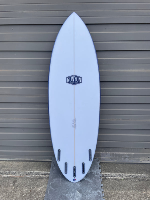 White surfboard leaning against sheet metal wall. Runyon Logo in the center. 4 +1 Future Fin Setup. Wider shortboard. 