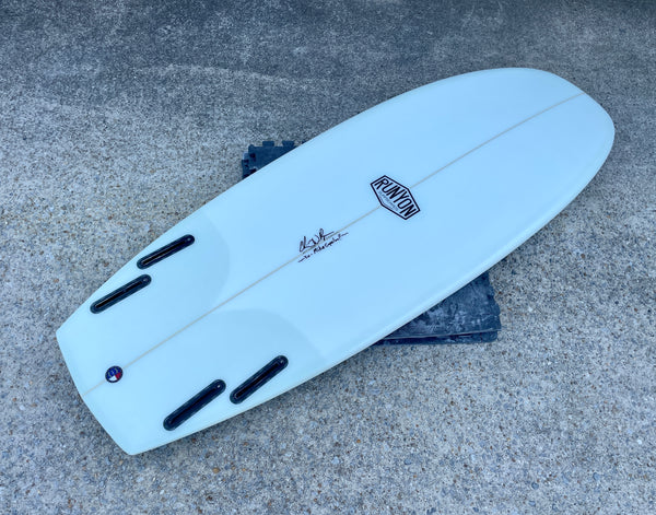 A white Mini hammer surfboard laying on its deck on a concrete. Quad fin setup. Runyon logo in the center. 