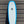 White Mini Hammer surfboard with light blue edges leaning against a sheet metal wall. Runyon Logo in the center. 