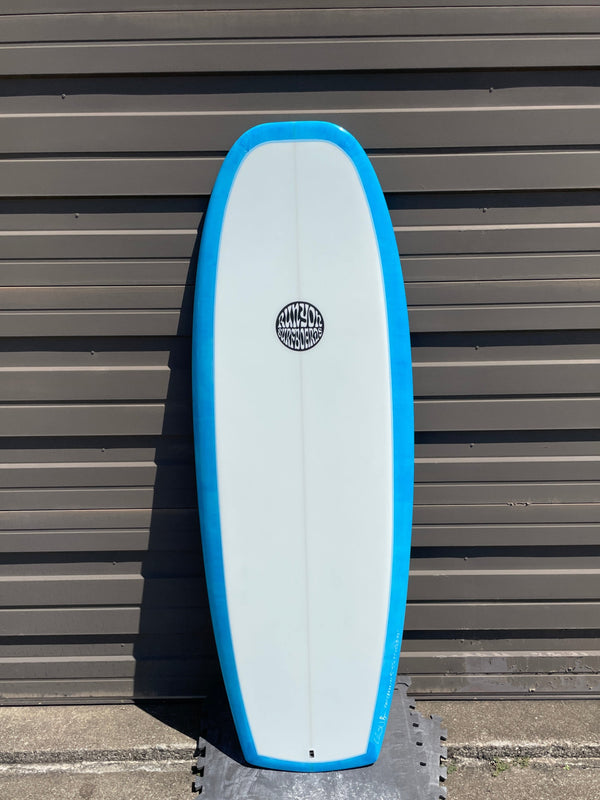 White Mini Hammer surfboard with light blue edges leaning against a sheet metal wall. Runyon Logo in the center. 