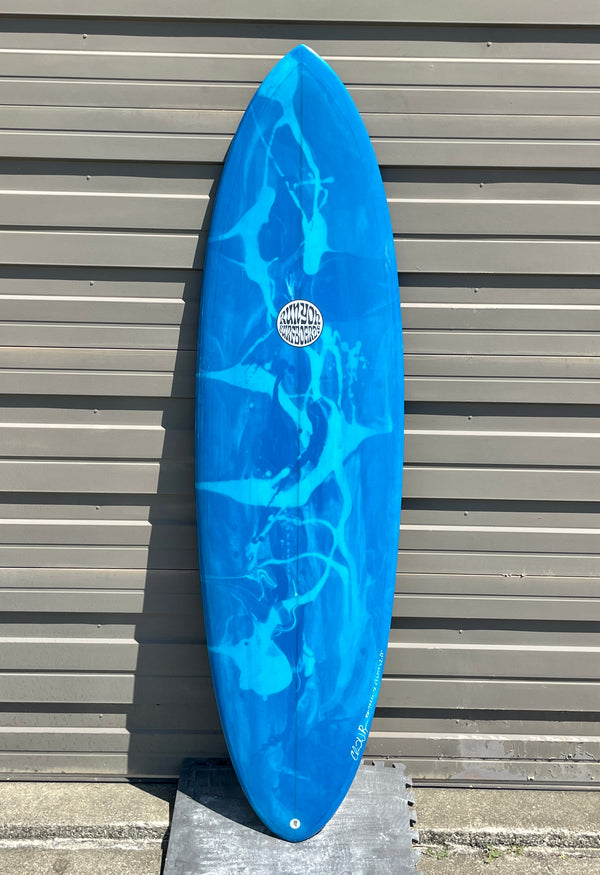 Blue Runyon "Dang Dang" Midlength Surfboard. Blue Marbling on Surfboard. Board is outside, leaning against a metal fabricate wall