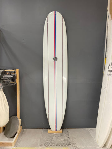 Classic White with Pinstripes Legend Longboard. Leaning against a gray wall. Red and Blue Striped run the length of the board. Runyon logo in the center. 