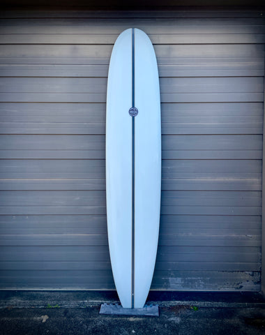A white surfboard with a logo leaning against a sheet metal wall.