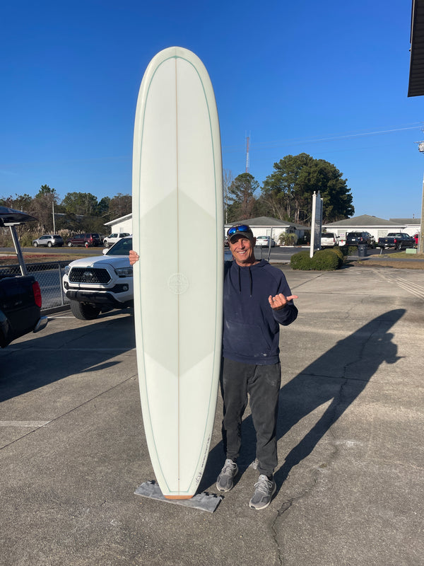 A man standing in a parking lot, showcasing a new longboard, that is standing on its end. 