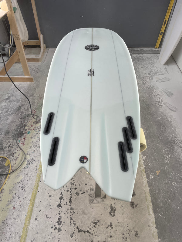 White asymmetric surfboard laying on stand in workshop. 3 Fin boxes on right side of fish tail, 2 fin boxes on the left. 