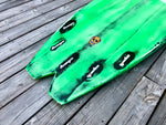 The image depicts a worn, green surfboard with a skull graphic on a wooden surface.