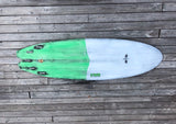 The image shows a colorful surfboard with a green and white design. The board has a skull graphic in the center and the letters "GWR" printed on the side. The board appears to be placed on a wooden surface, suggesting it is resting on a deck or pier.